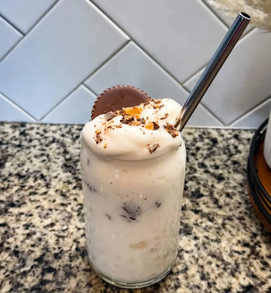 Finished Ninja SLUSHi Peanut Butter Cup Vanilla Protein Shake in a glass, topped with whipped cream, crushed peanut butter cup pieces, and a whole peanut butter cup, with a metal straw for serving.