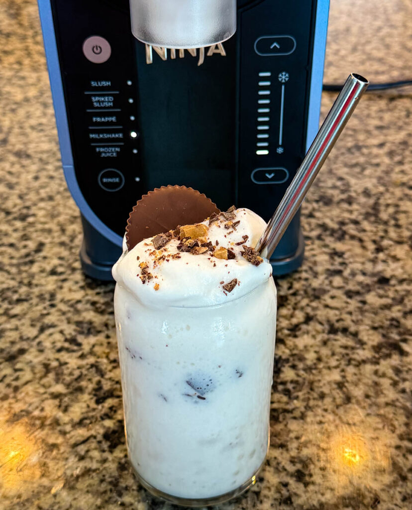 Finished Ninja SLUSHi Peanut Butter Cup Vanilla Protein Shake in a glass, topped with whipped cream, crushed peanut butter cup pieces, and a whole peanut butter cup, with a metal straw for serving.