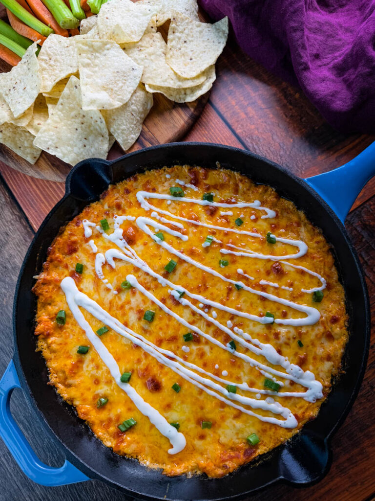 Hot and cheesy Buffalo Chicken Dip in a black cast-iron skillet, garnished with fresh green onions and a drizzle of ranch dressing, ready to be served.