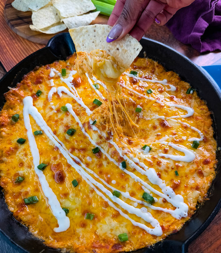 Golden, bubbly Ranch Buffalo Chicken Dip with Rotisserie Chicken in a skillet, topped with drizzled ranch dressing and chopped green onions. A tortilla chip is being dipped, showing the gooey, cheesy pull.