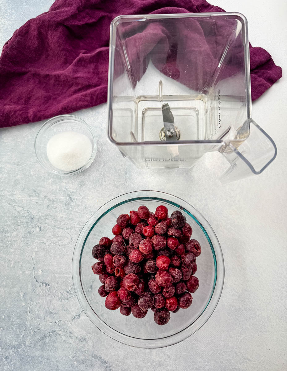 A flat-lay image displaying a clear blender jar, a bowl of frozen cherries, and a small glass bowl of sugar, ready to be blended into a cherry slushi. The background has a neutral textured surface with a deep purple cloth.