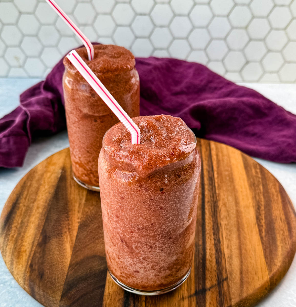 A close-up shot of two frosty cherry slushies with a rich red color and perfectly blended texture. The wooden board and purple cloth create a cozy, inviting atmosphere.