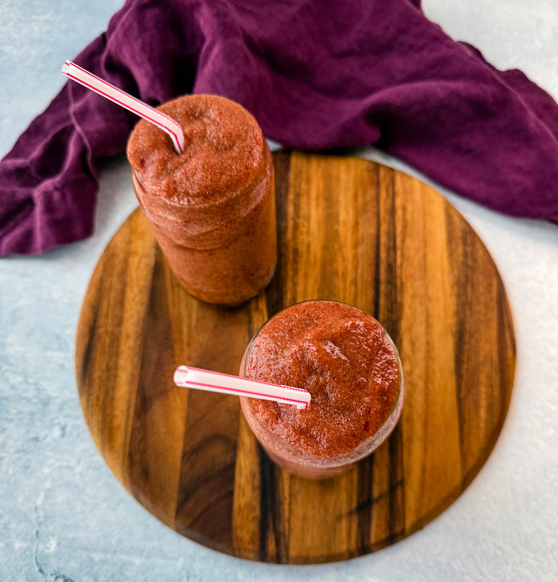 An overhead view of two tall glasses filled with frozen cherry slushies, placed on a wooden serving board. The drinks are garnished with striped straws, and a purple cloth adds contrast in the background.