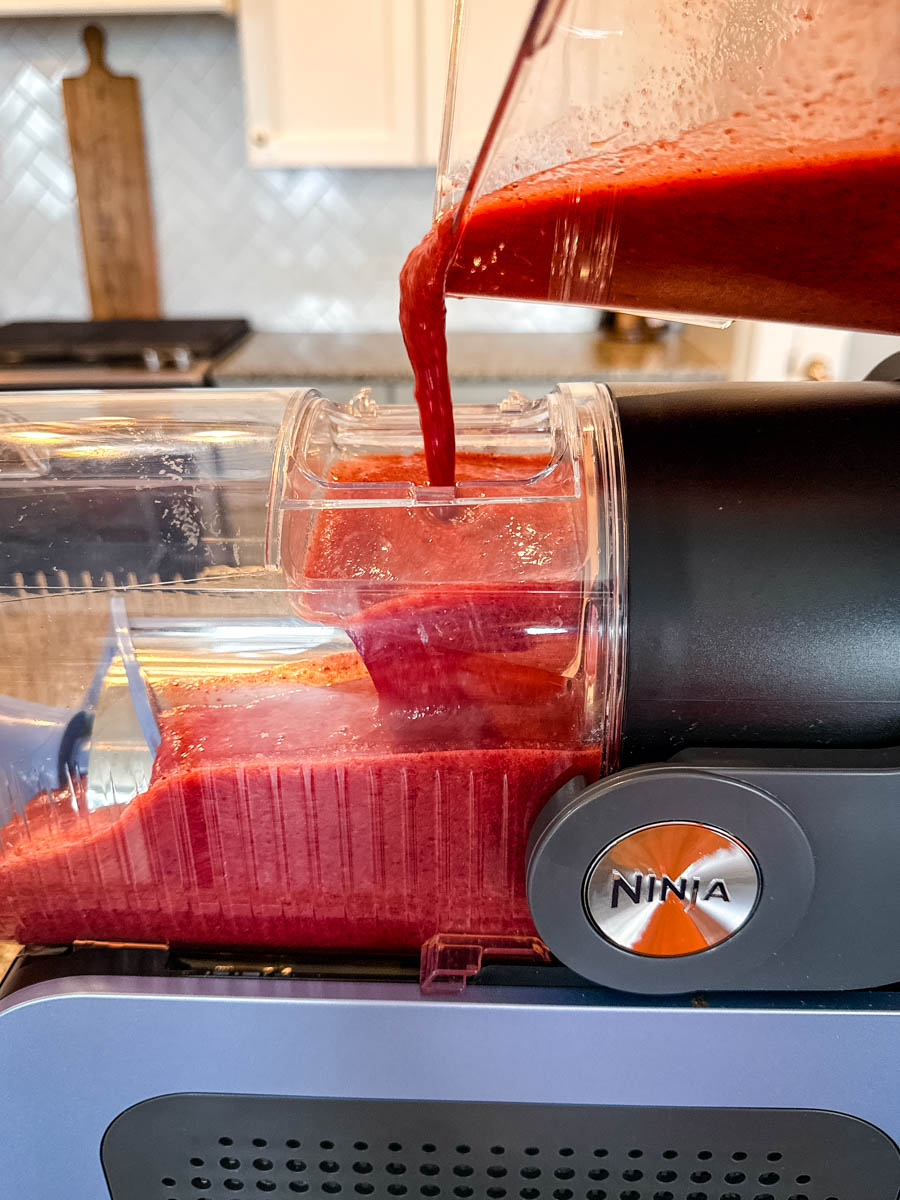 A close-up of a vibrant red cherry mixture being poured from a blender into the Ninja Slushi Machine. The background features a bright kitchen with a white backsplash and wooden cutting board.