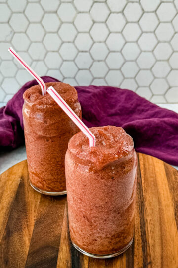 Two tall glasses filled with thick, icy cherry slushies, served with red-and-white striped straws. The drinks are placed on a round wooden serving board, with a deep purple cloth draped in the background against a white hexagonal tile backdrop.