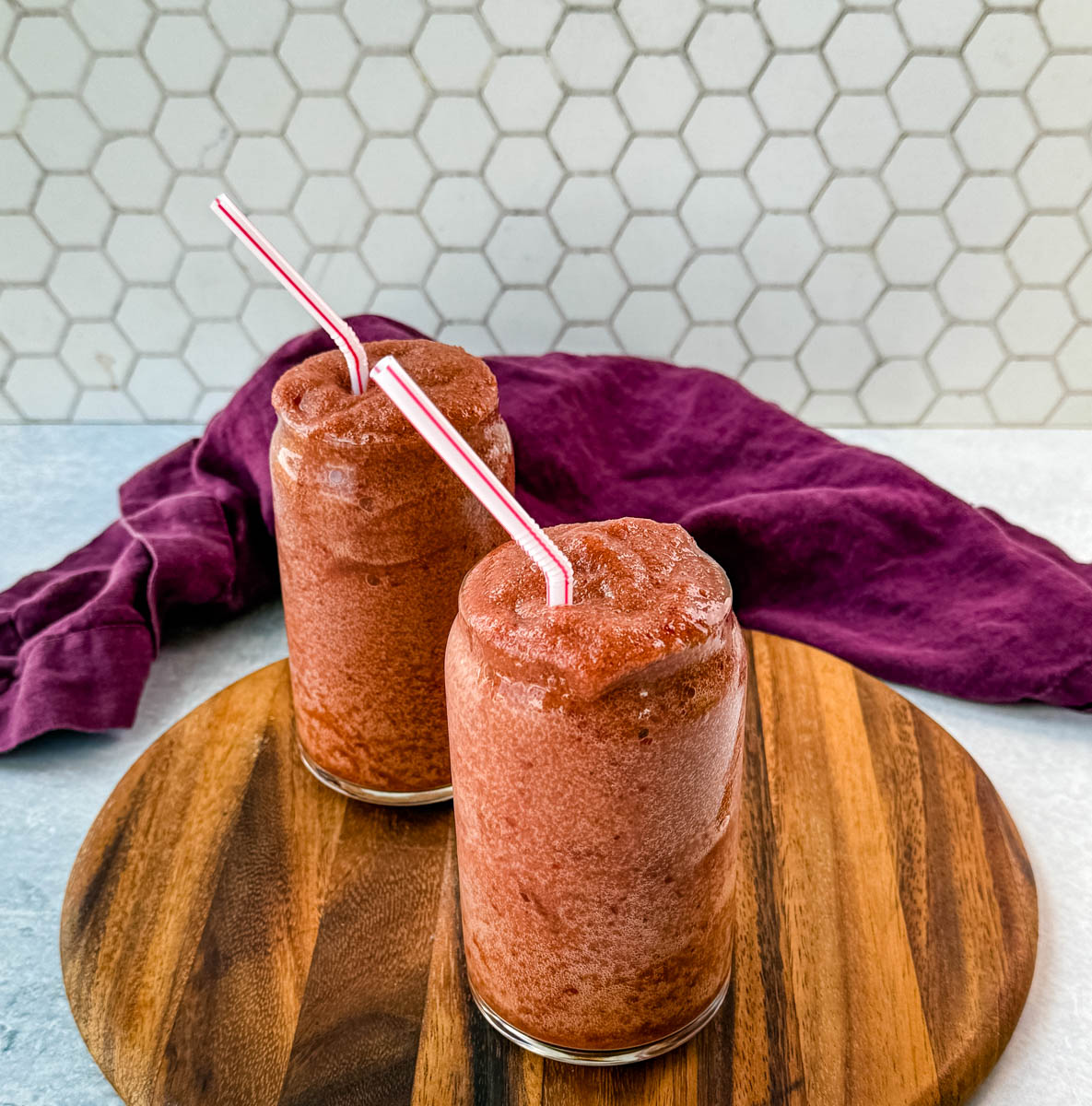 Two tall glasses filled with thick, icy cherry slushies, served with red-and-white striped straws. The drinks are placed on a round wooden serving board, with a deep purple cloth draped in the background against a white hexagonal tile backdrop.