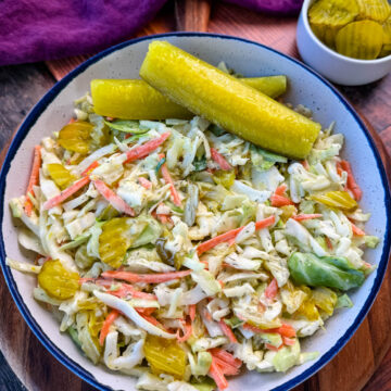 Bowl of Dill Pickle Coleslaw with Pickle Spears on the Side – A close-up of a creamy dill pickle coleslaw served in a rustic bowl, featuring shredded cabbage, carrots, and sliced pickles, garnished with two large dill pickle spears. A small dish of extra pickle slices sits in the background on a wooden surface.