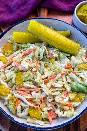 Bowl of Dill Pickle Coleslaw with Pickle Spears on the Side – A close-up of a creamy dill pickle coleslaw served in a rustic bowl, featuring shredded cabbage, carrots, and sliced pickles, garnished with two large dill pickle spears. A small dish of extra pickle slices sits in the background on a wooden surface.