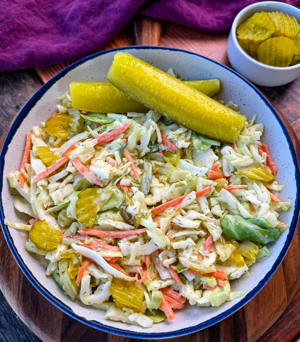 Bowl of Dill Pickle Coleslaw with Pickle Spears on the Side – A close-up of a creamy dill pickle coleslaw served in a rustic bowl, featuring shredded cabbage, carrots, and sliced pickles, garnished with two large dill pickle spears. A small dish of extra pickle slices sits in the background on a wooden surface.