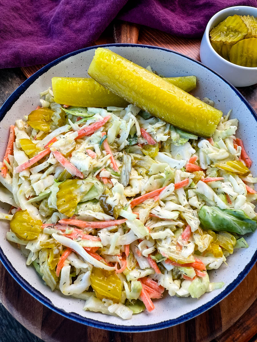 Bowl of Dill Pickle Coleslaw with Pickle Spears on the Side – A close-up of a creamy dill pickle coleslaw served in a rustic bowl, featuring shredded cabbage, carrots, and sliced pickles, garnished with two large dill pickle spears. A small dish of extra pickle slices sits in the background on a wooden surface.