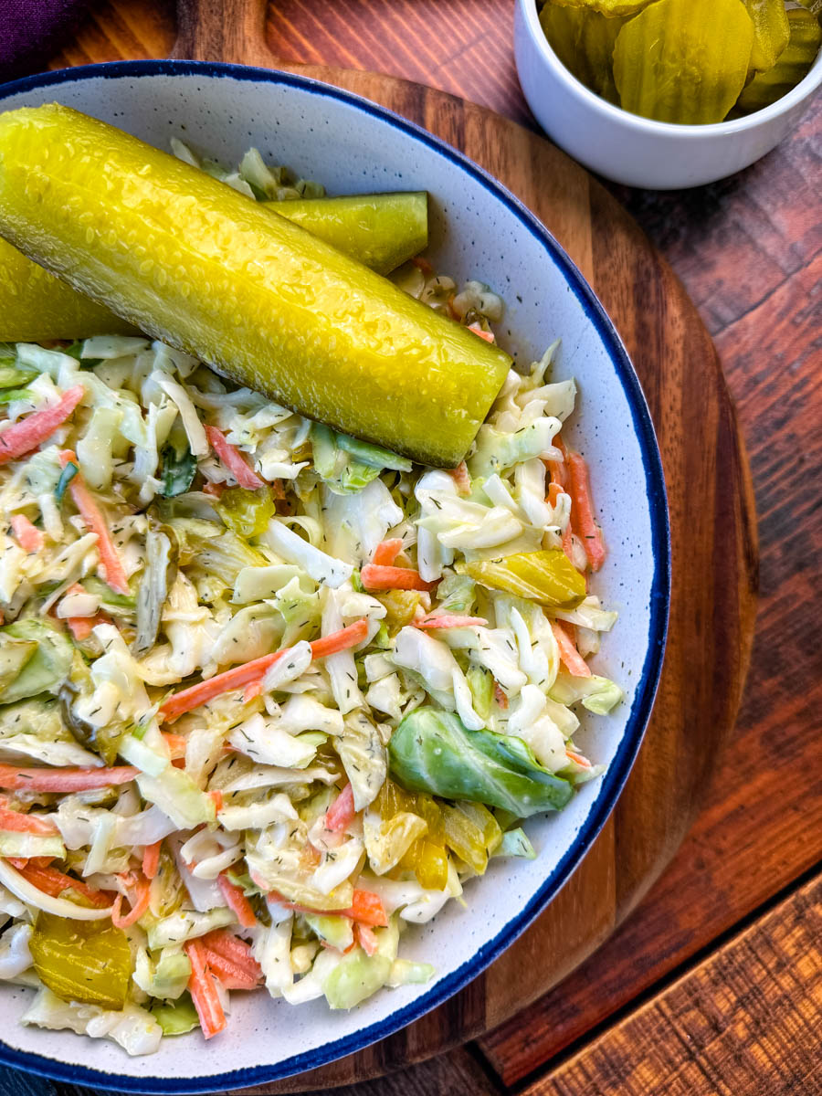 Bowl of Dill Pickle Coleslaw with Pickle Spears on the Side – A close-up of a creamy dill pickle coleslaw served in a rustic bowl, featuring shredded cabbage, carrots, and sliced pickles, garnished with two large dill pickle spears. A small dish of extra pickle slices sits in the background on a wooden surface.