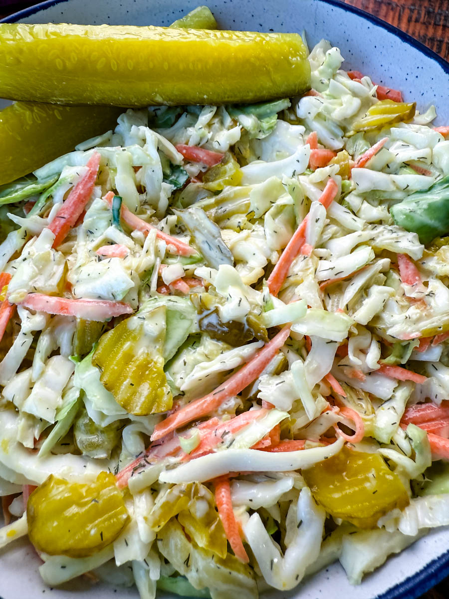 Bowl of Dill Pickle Coleslaw with Pickle Spears on the Side – A close-up of a creamy dill pickle coleslaw served in a rustic bowl, featuring shredded cabbage, carrots, and sliced pickles, garnished with two large dill pickle spears. A small dish of extra pickle slices sits in the background on a wooden surface.