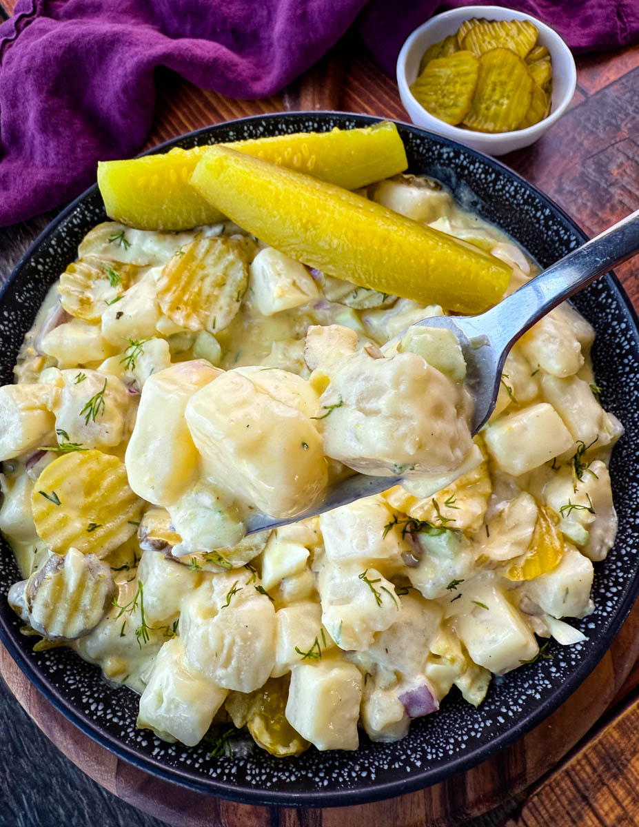 A spoon lifting a bite of creamy dill pickle potato salad, showcasing the texture of the cubed potatoes, crunchy pickles, and fresh dill. The bowl of salad sits on a wooden surface with a small dish of pickles in the background, creating a visually appetizing presentation.