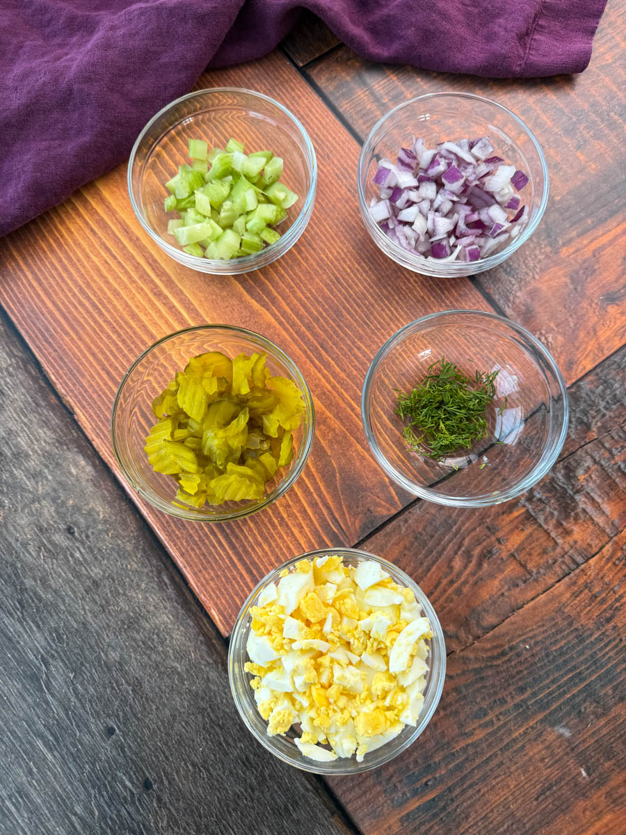 A top-down view of five small glass bowls containing prepped ingredients for dill pickle potato salad. The bowls hold diced celery, chopped red onion, chopped dill pickles, fresh dill, and chopped hard-boiled eggs. They are placed on a wooden surface with a dark purple cloth in the background.