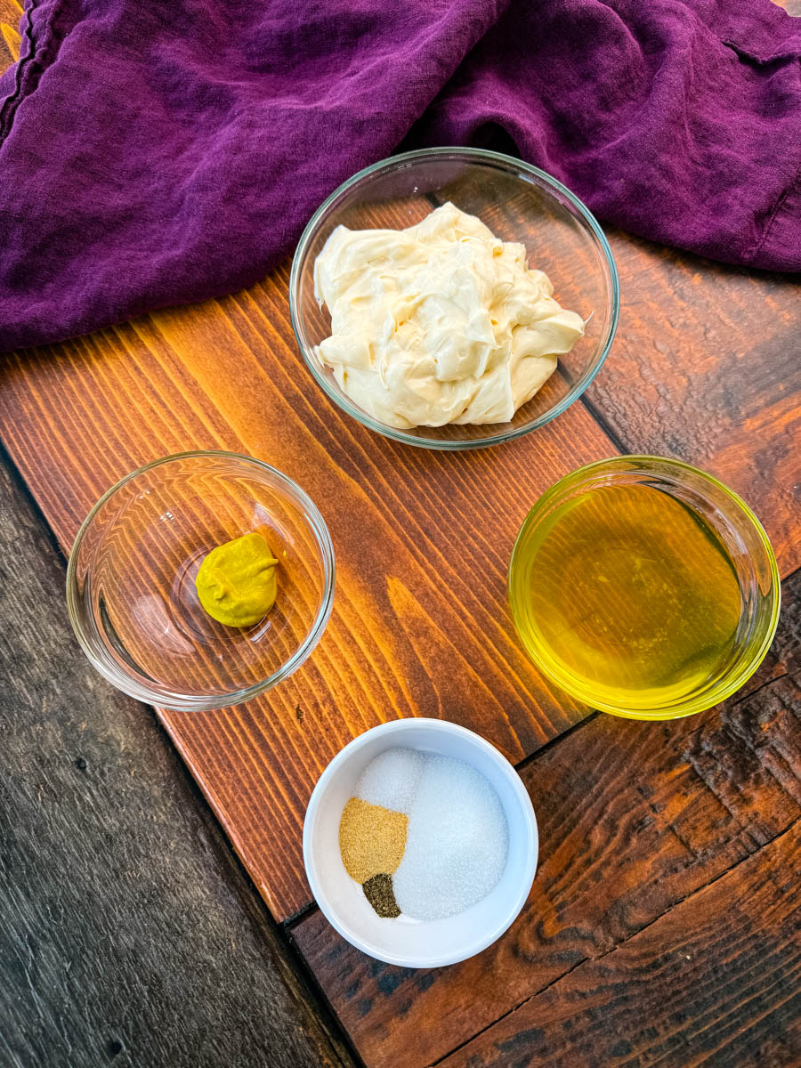 A flat-lay image displaying small glass bowls with ingredients for the salad dressing. The bowls contain mayonnaise, yellow mustard, pickle juice, and a blend of seasonings including salt, pepper, and garlic powder. The wooden surface and purple cloth create a rustic background.