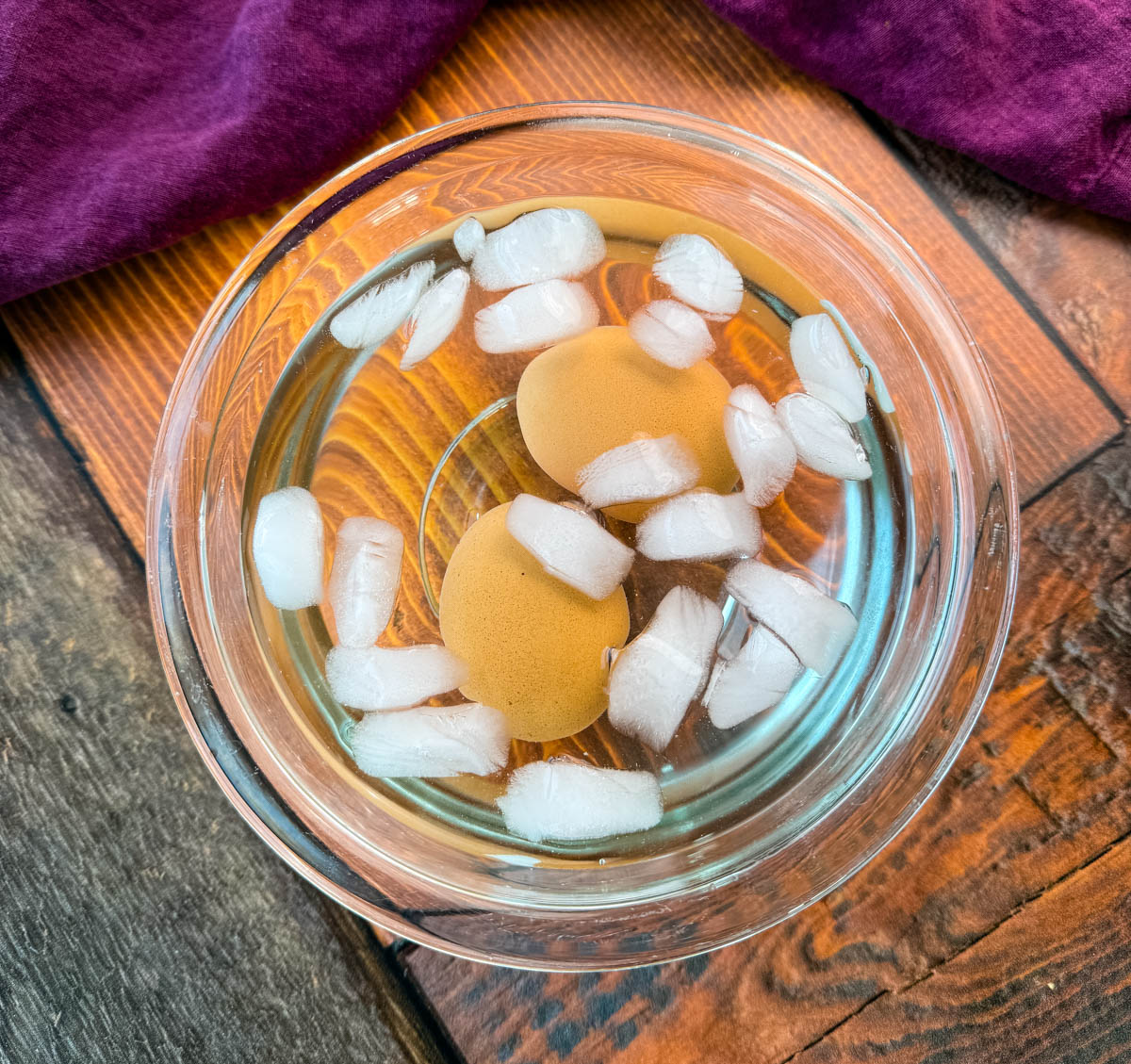 A close-up of two brown eggs submerged in a glass bowl filled with ice water. Ice cubes float in the water, helping to cool the eggs quickly after boiling. The bowl sits on a wooden surface with a deep purple cloth in the background.