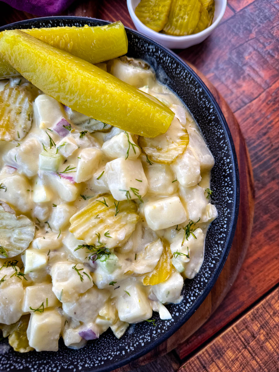 A close-up image of a bowl of creamy dill pickle potato salad. The salad features cubed potatoes, diced pickles, red onion, celery, and fresh dill, all coated in a creamy dressing. The dish is garnished with sliced pickles and served in a black speckled bowl, with a background of a purple cloth and a small dish of pickles.