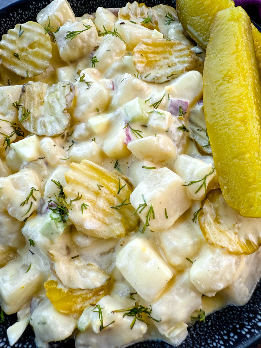 A close-up image of a bowl of creamy dill pickle potato salad. The salad features cubed potatoes, diced pickles, red onion, celery, and fresh dill, all coated in a creamy dressing. The dish is garnished with sliced pickles and served in a black speckled bowl, with a background of a purple cloth and a small dish of pickles.