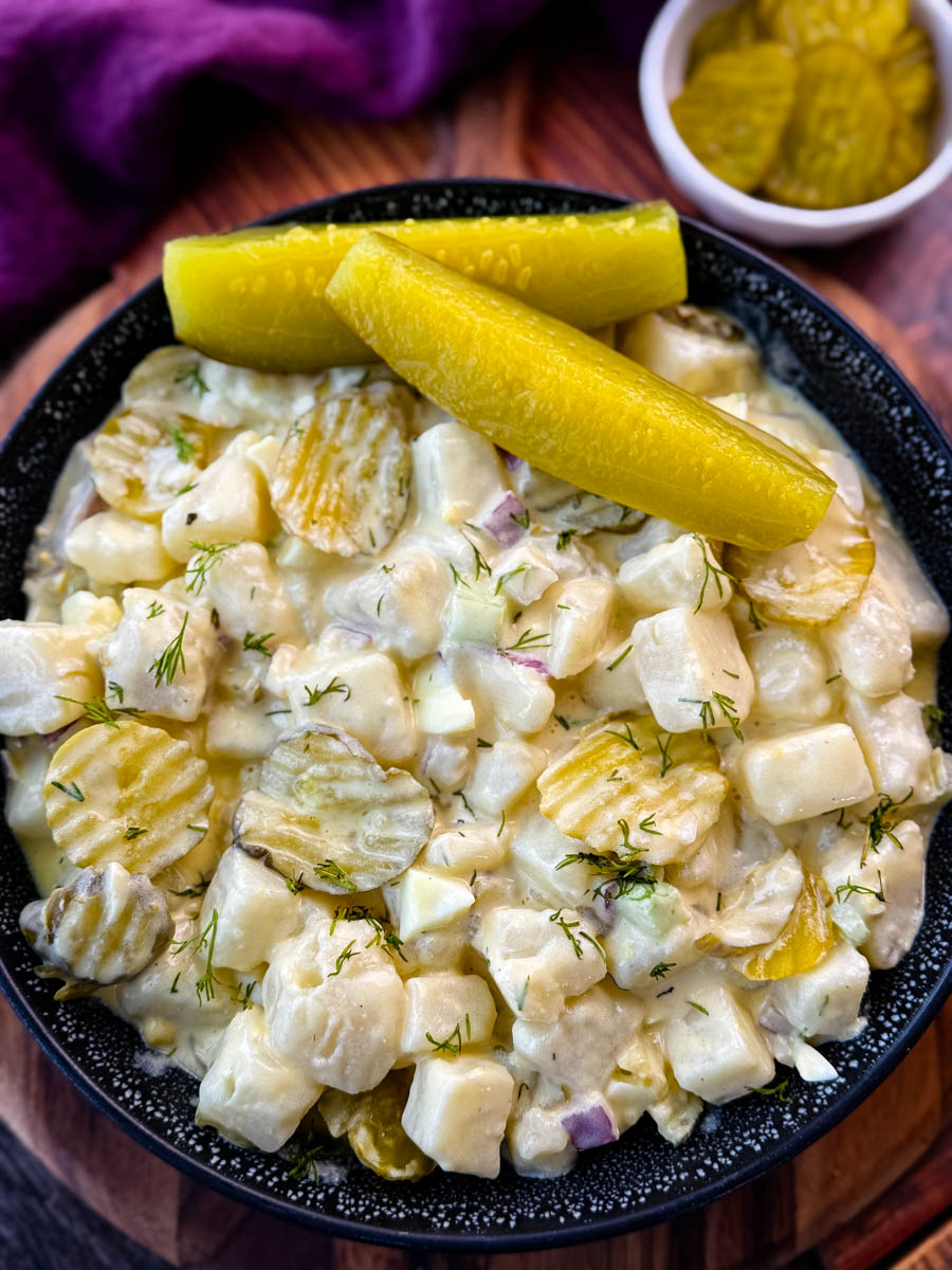 A close-up image of a bowl of creamy dill pickle potato salad. The salad features cubed potatoes, diced pickles, red onion, celery, and fresh dill, all coated in a creamy dressing. The dish is garnished with sliced pickles and served in a black speckled bowl, with a background of a purple cloth and a small dish of pickles.