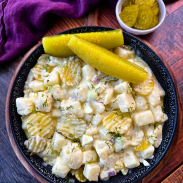 A close-up image of a bowl of creamy dill pickle potato salad. The salad features cubed potatoes, diced pickles, red onion, celery, and fresh dill, all coated in a creamy dressing. The dish is garnished with sliced pickles and served in a black speckled bowl, with a background of a purple cloth and a small dish of pickles.