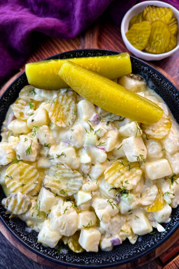 A close-up image of a bowl of creamy dill pickle potato salad. The salad features cubed potatoes, diced pickles, red onion, celery, and fresh dill, all coated in a creamy dressing. The dish is garnished with sliced pickles and served in a black speckled bowl, with a background of a purple cloth and a small dish of pickles.