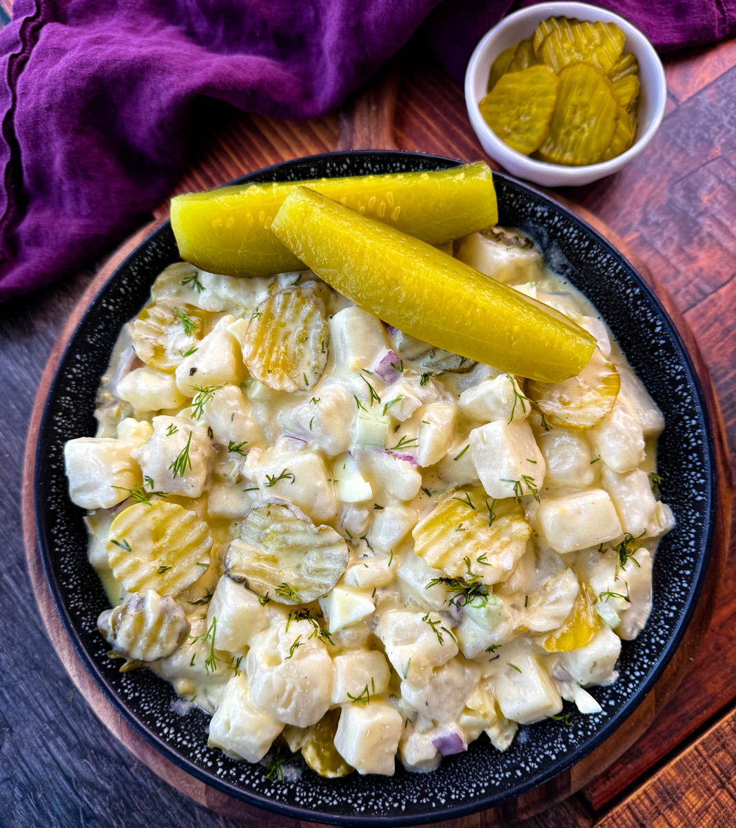 A close-up image of a bowl of creamy dill pickle potato salad. The salad features cubed potatoes, diced pickles, red onion, celery, and fresh dill, all coated in a creamy dressing. The dish is garnished with sliced pickles and served in a black speckled bowl, with a background of a purple cloth and a small dish of pickles.