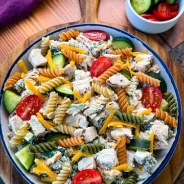 Final Plated Chicken Pasta Salad with Side Dish of Extra Vegetables – A full view of the pasta salad served in a bowl with a small dish of extra cherry tomatoes and cucumbers on the side, placed on a rustic wooden board.