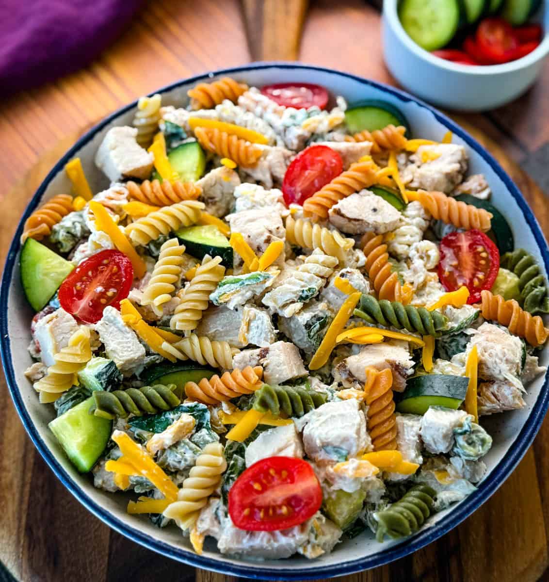 Overhead View of Chicken Pasta Salad in a Bowl – A beautifully plated pasta salad with a mix of textures and colors, including red cherry tomatoes, green cucumbers, and shredded cheddar cheese, served in a rustic dish on a wooden surface.