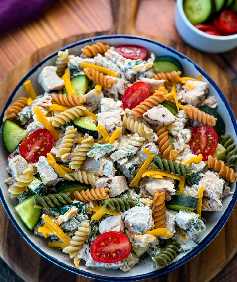 Overhead View of Chicken Pasta Salad in a Bowl – A beautifully plated pasta salad with a mix of textures and colors, including red cherry tomatoes, green cucumbers, and shredded cheddar cheese, served in a rustic dish on a wooden surface.