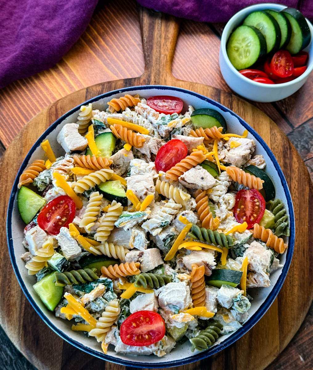 Overhead View of Chicken Pasta Salad in a Bowl – A beautifully plated pasta salad with a mix of textures and colors, including red cherry tomatoes, green cucumbers, and shredded cheddar cheese, served in a rustic dish on a wooden surface.