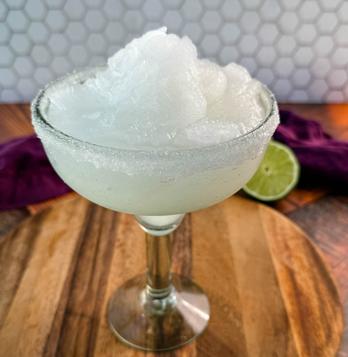 A close-up of a margarita glass filled with frozen slush, highlighting the fluffy, snow-like texture of the drink. The sugar-rimmed glass sits on a rustic wooden board.