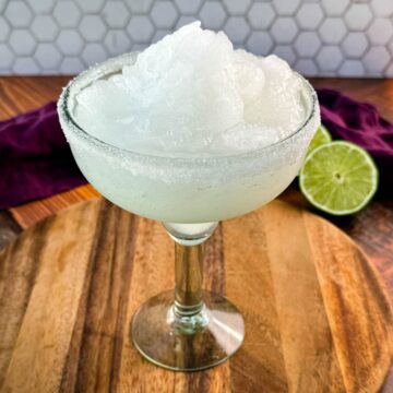 A close-up of a margarita glass filled with frozen slush, highlighting the fluffy, snow-like texture of the drink. The sugar-rimmed glass sits on a rustic wooden board.