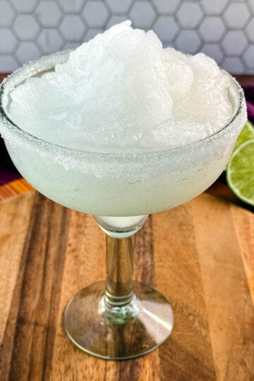 A close-up of a margarita glass filled with frozen slush, highlighting the fluffy, snow-like texture of the drink. The sugar-rimmed glass sits on a rustic wooden board.