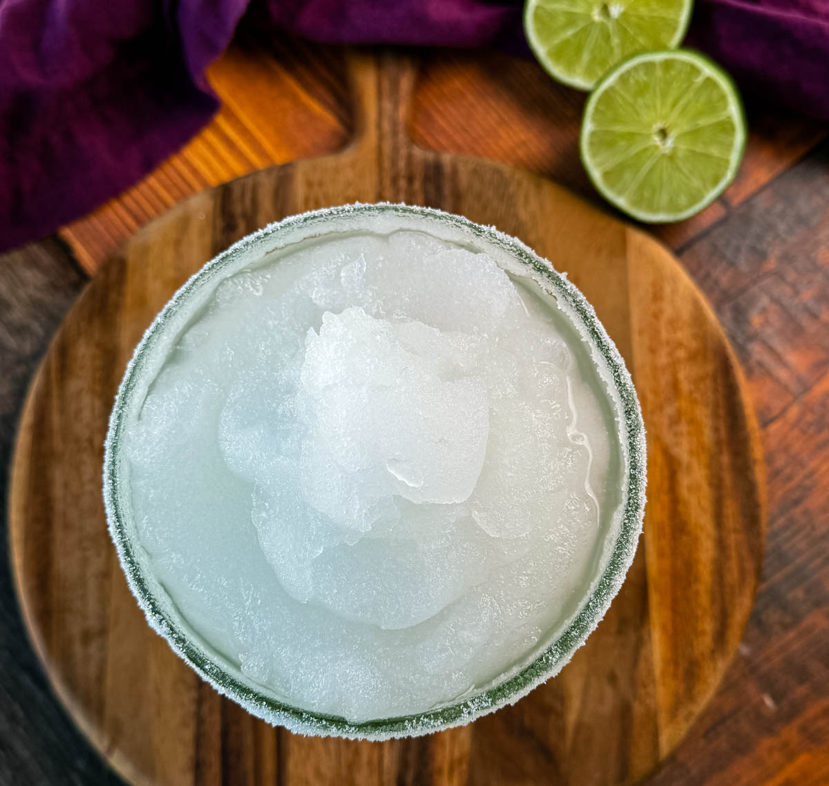 A top-down shot of a frozen margarita in a glass with a sugar rim, showcasing the smooth and icy texture. Fresh lime slices and a purple cloth napkin are visible in the background.