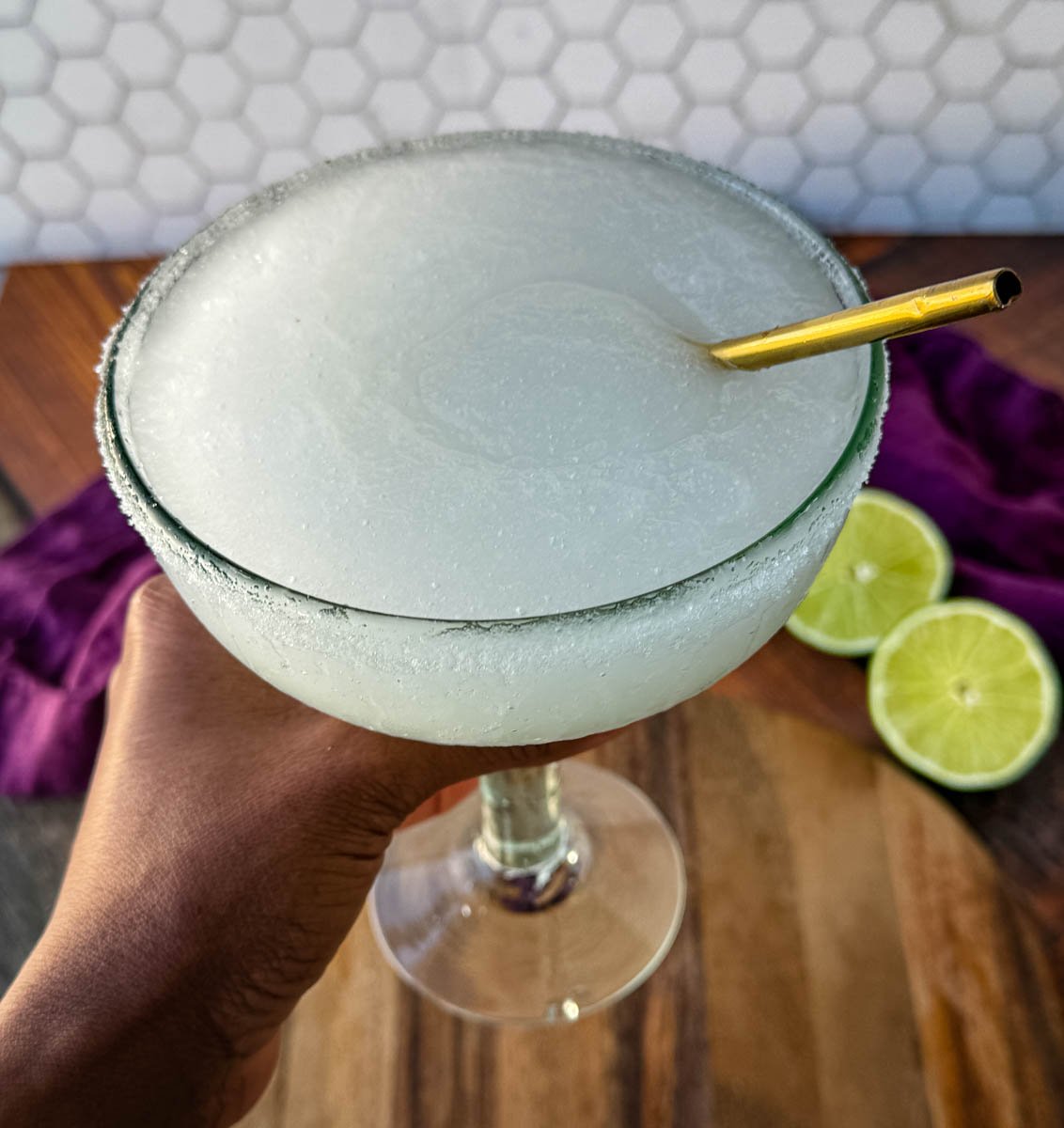 A hand holding a large margarita glass with a sugar rim, filled with a classic frozen margarita, with a golden straw inserted. Lime slices and a purple napkin sit in the background.