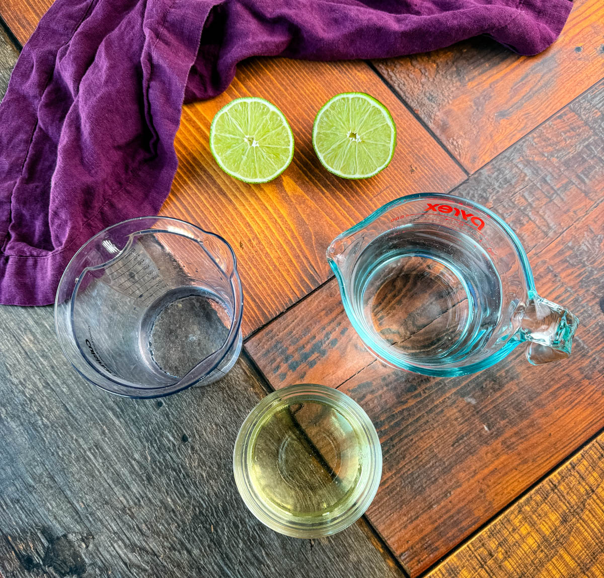 A flat lay of margarita ingredients including a measuring cup of water, lime halves, and small bowls of simple syrup and tequila, displayed on a wooden table with a dark purple napkin.