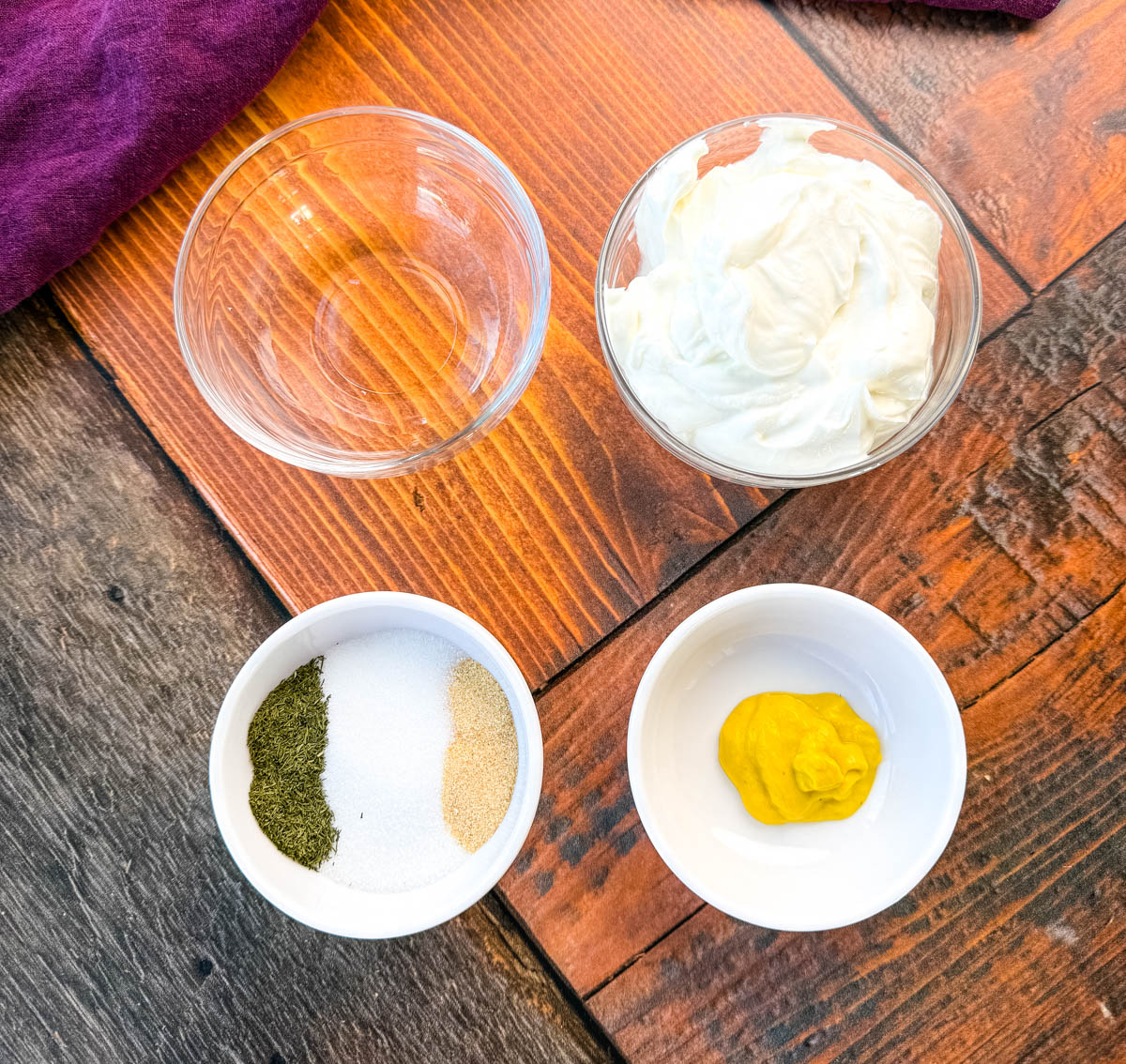 Coleslaw Dressing Ingredients in Small Bowls – A top-down view of individual ingredients in small white bowls on a rustic wooden surface, including Greek yogurt, mustard, dried dill, sugar, garlic powder, and an empty bowl for pickle juice. A dark purple cloth is partially visible in the background.