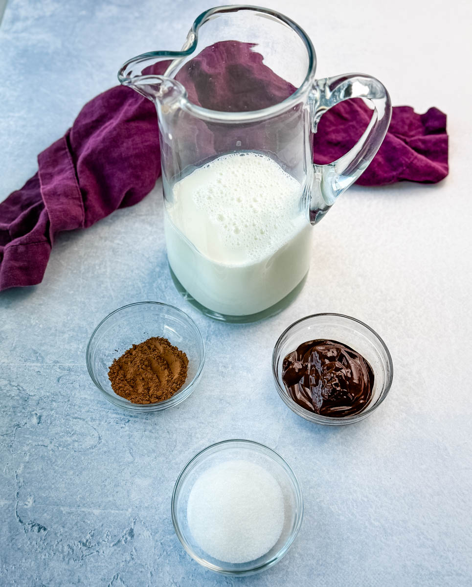 A glass pitcher filled with milk, surrounded by small glass bowls containing cocoa powder, granulated sugar, and melted chocolate, set on a light blue textured surface with a purple cloth in the background.