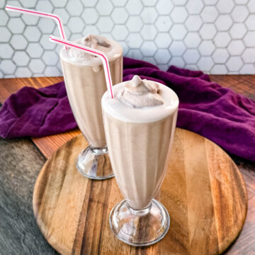 Two glasses of homemade Wendy’s Frosty-style milkshakes served with pink-striped straws, placed on a wooden board with a purple cloth in the background and a hexagonal tile backsplash.