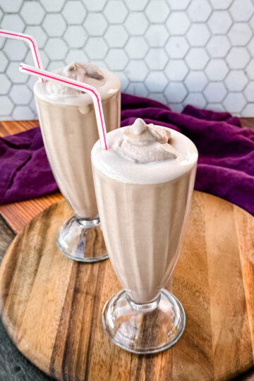 Two glasses of homemade Wendy’s Frosty-style milkshakes served with pink-striped straws, placed on a wooden board with a purple cloth in the background and a hexagonal tile backsplash.