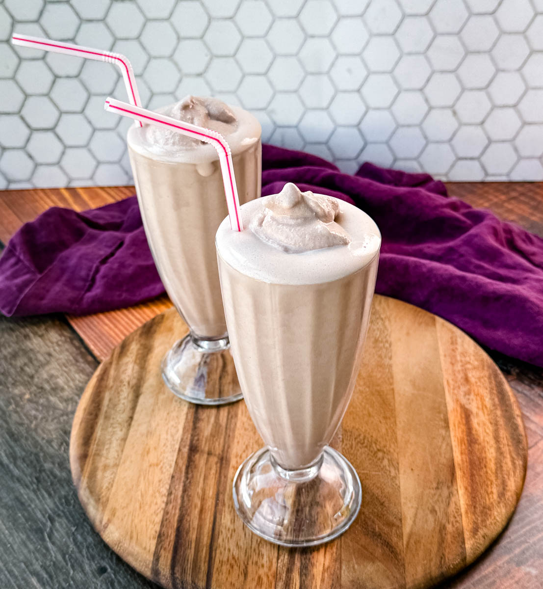 Two glasses of homemade Wendy’s Frosty-style milkshakes served with pink-striped straws, placed on a wooden board with a purple cloth in the background and a hexagonal tile backsplash.