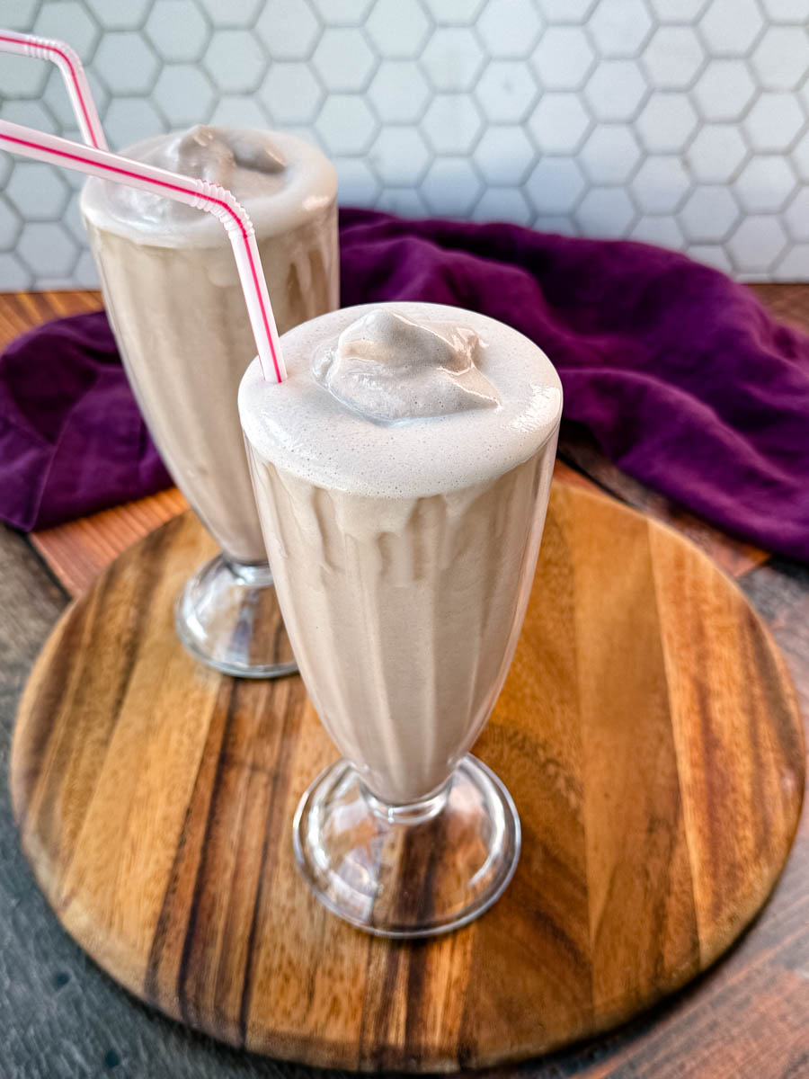 Two glasses of homemade Wendy’s Frosty-style milkshakes served with pink-striped straws, placed on a wooden board with a purple cloth in the background and a hexagonal tile backsplash.