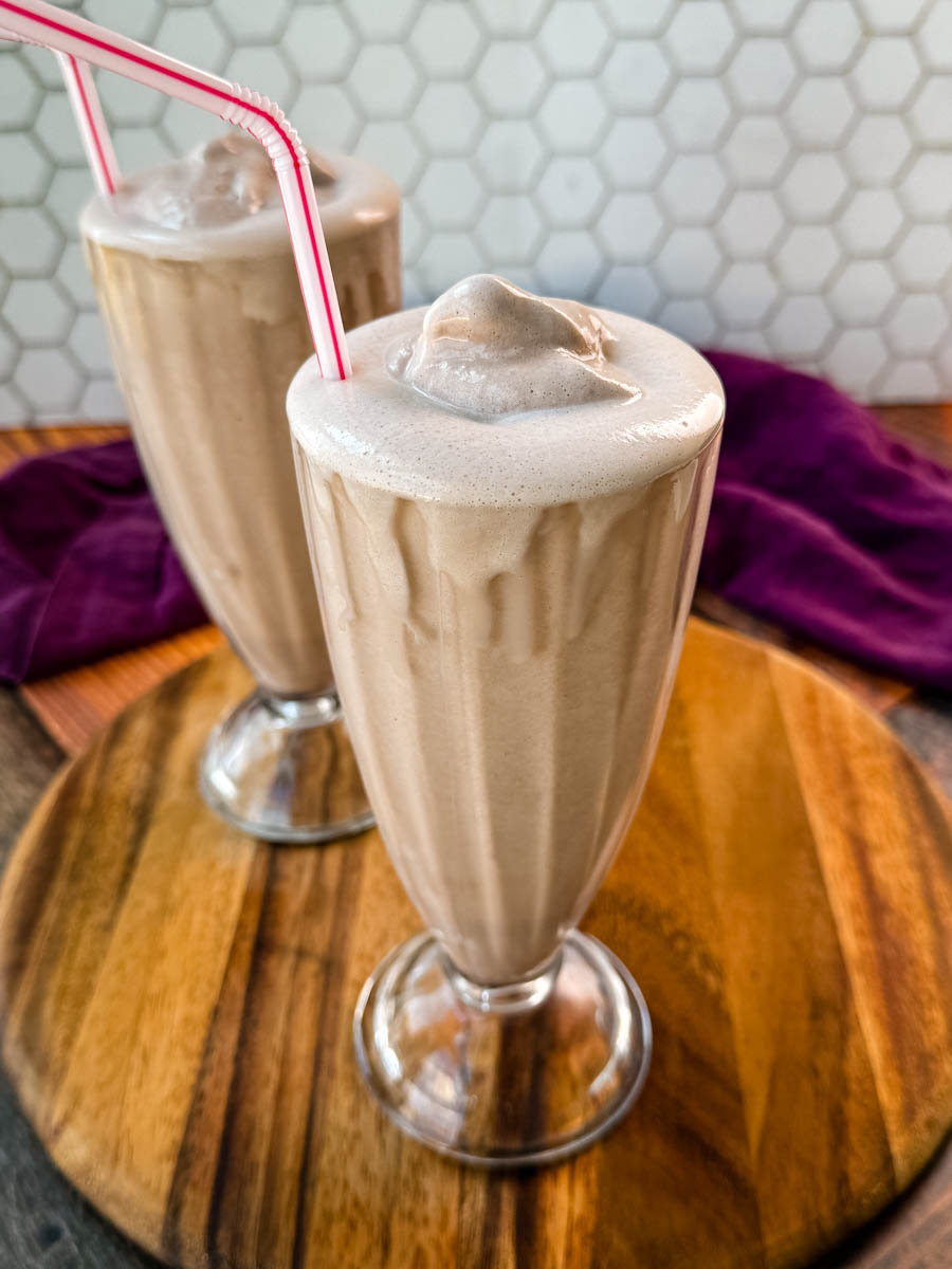 Two glasses of homemade Wendy’s Frosty-style milkshakes served with pink-striped straws, placed on a wooden board with a purple cloth in the background and a hexagonal tile backsplash.