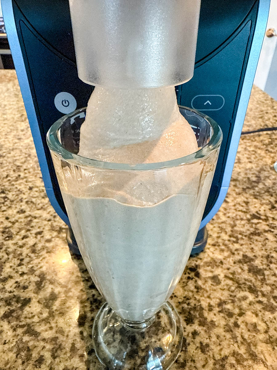 A thick, creamy Wendy’s Frosty-style milkshake being dispensed from a Ninja slushie machine into a tall glass on a granite countertop.